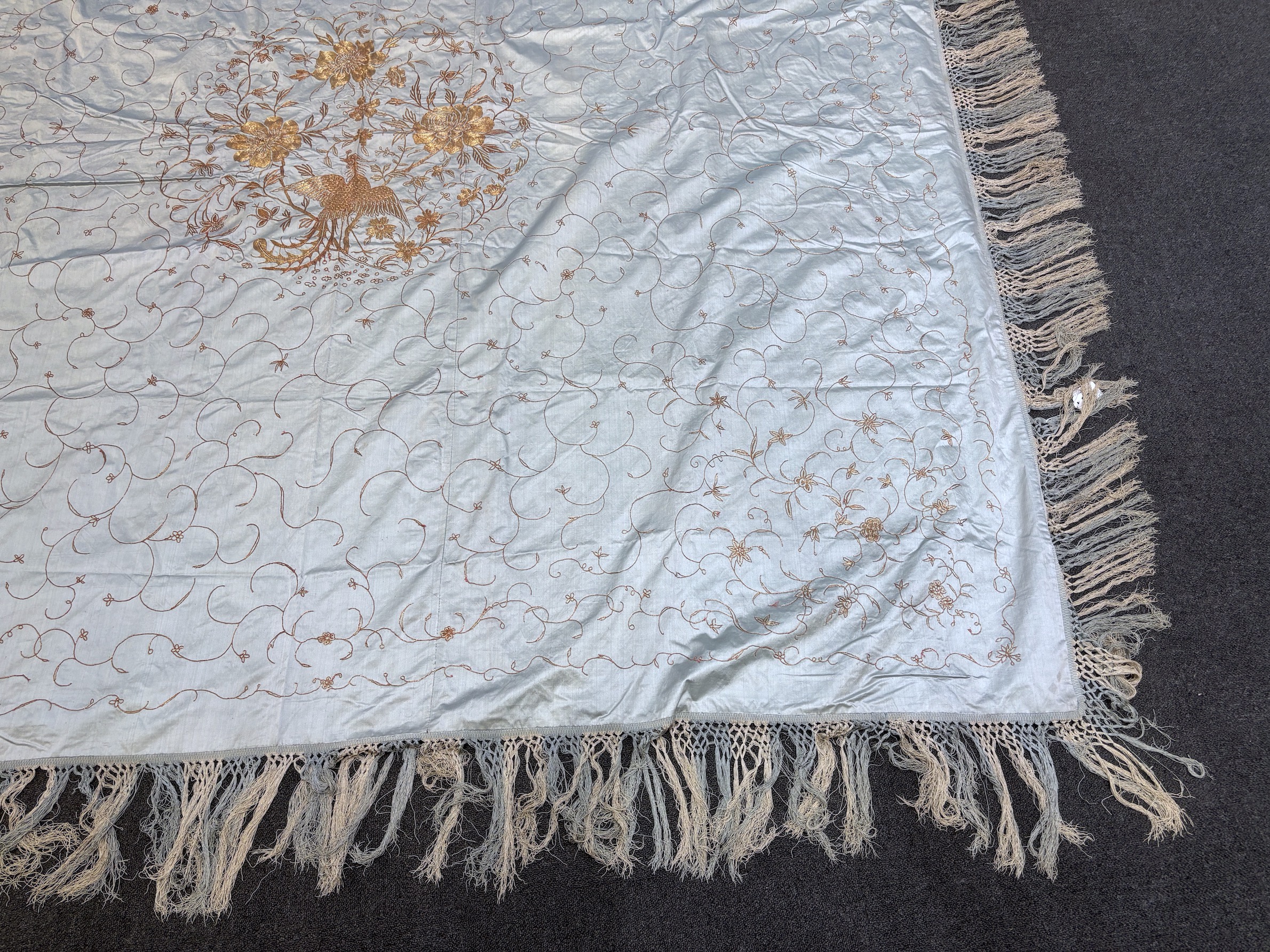 A Chinese late 19th/early 20th century silk satin pale blue bed cover, embroidered with gold thread and a central motif of a phoenix, flowers and leaves, bordered with cream and blue fringing on three sides and backed wi